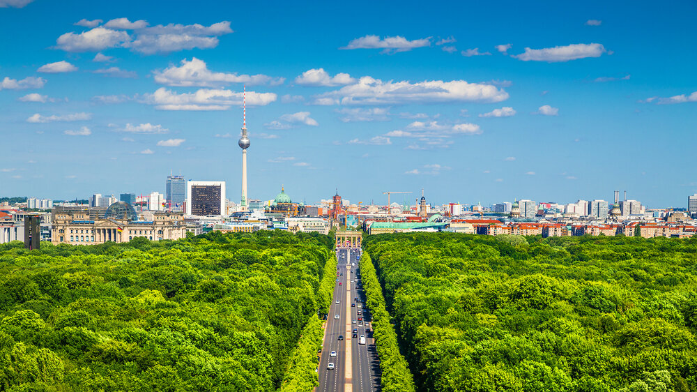 Panorama von Berlin mit Blick über das grüne Tiergartenviertel, im Hintergrund der Fernsehturm, das Brandenburger Tor und die Skyline bei blauem Himmel.