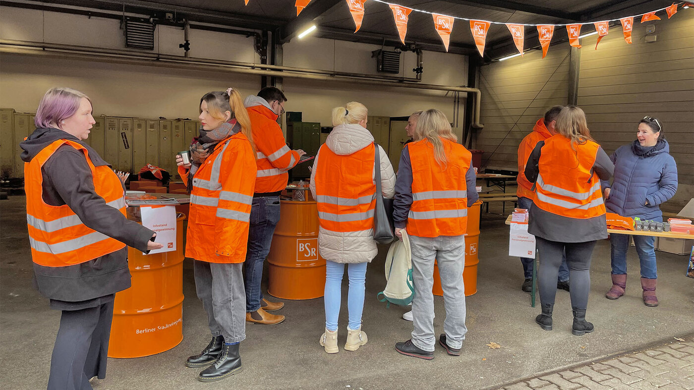 Eine Gruppe von Menschen in Warnwesten steht in einer Halle mit Infoständen. Auf den Tischen liegt Info-Material, mehrere Personen sind im Gespräch.