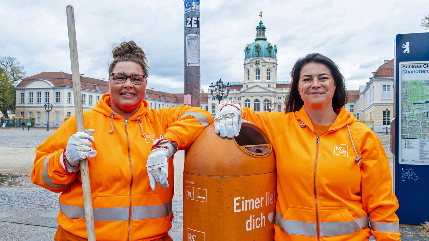 Zwei BSR-Mitarbeiterinnen in orangefarbener Arbeitskleidung stehen im urbanen Raum neben einem Mülleimer mit der Aufschrift „Eimer liebt dich“.