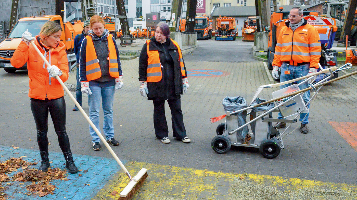 Eine Gruppe von Menschen in Warnwesten steht auf dem Hof der BSR. Im Vordergrund fegt eine Frau. Im Hintergrund sind BSR-Fahrzeuge zu sehen.