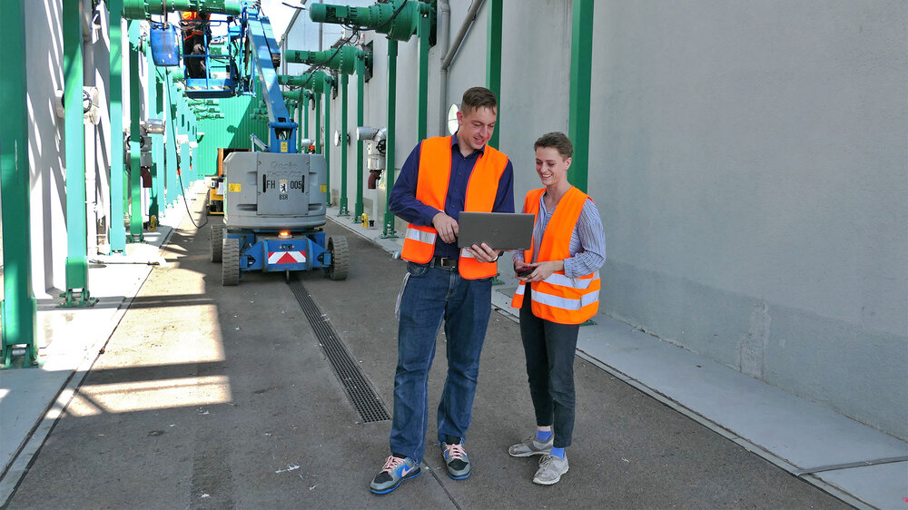 Zwei Personen in orangen Sicherheitswesten stehen in einem Industrieumfeld und schauen auf einen Laptop.