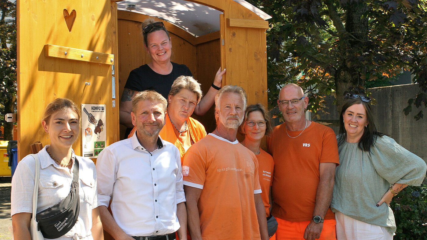 Eine Gruppe von Menschen und drei BSR-Mitarbeitende stehen vor einer Komposttoilette aus Holz.
