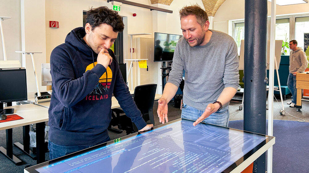 Zwei Männer stehen an einem Touchscreen-Tisch, auf dem Programmcode angezeigt wird. Einer gestikuliert, der andere steht in nachdenklicher Pose.