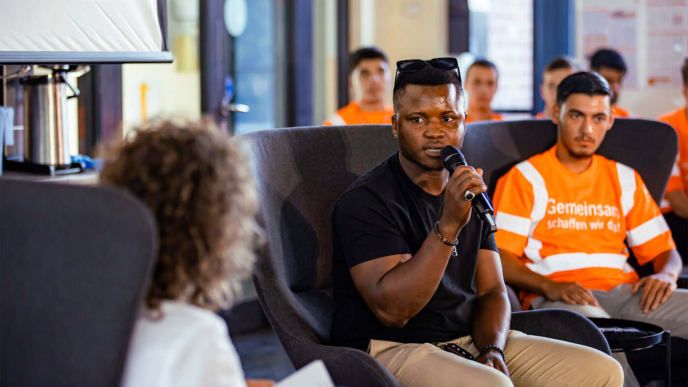 Ein junger Mann mit Sonnenbrille auf dem Kopf sitzt in einem Sessel und spricht in ein Mikrofon, während andere in orangefarbenen Shirts im Hintergrund sitzen.