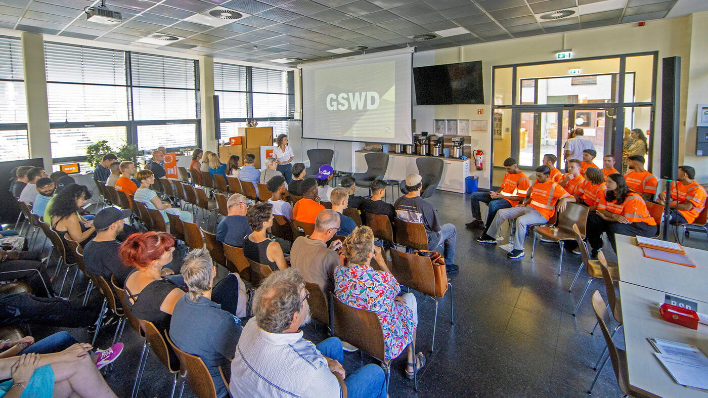 Ein großer Raum mit vielen Stühlen, auf denen Menschen sitzen. Vorne steht eine Leinwand mit der Aufschrift „GSWD“. Einige der sitzenden Personen tragen orangefarbene T-Shirts.