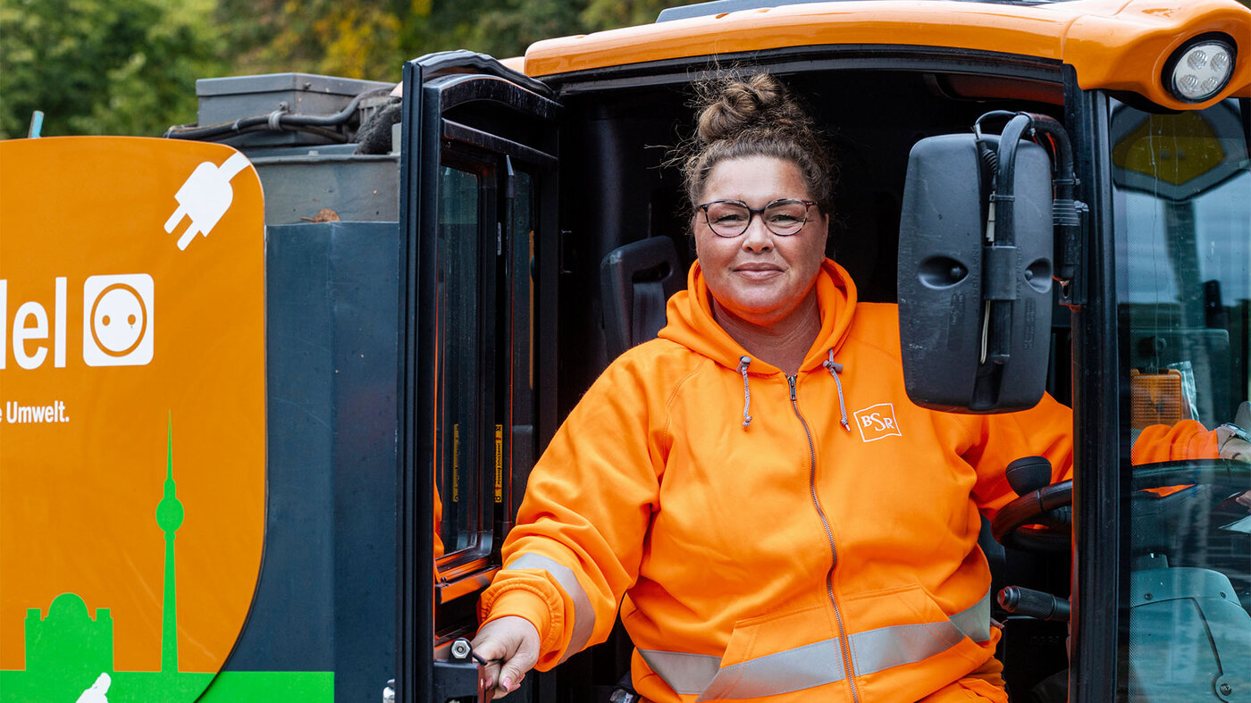 Eine Frau mit Brille und Dutt sitzt lächelnd am Steuer eines orangefarbenen Straßenkehrfahrzeuges. Sie trägt orangefarbene Arbeitskleidung.
