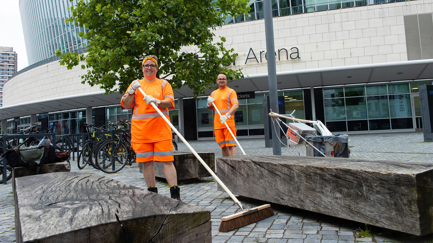 Zwei lächelnde Personen in orangefarbener Arbeitskleidung und Handschuhen halten Besen und stehen vor dem Arena-Gebäude. 