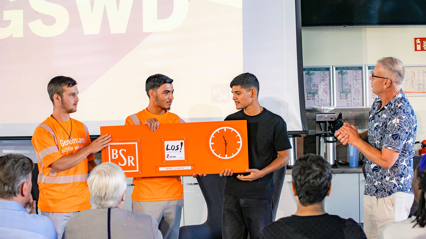 Drei junge Männer in orangefarbenen Shirts halten gemeinsam ein großes orangefarbenes Schild mit den Aufschriften „BSR“, „LOS!“ und einer Uhr in die Höhe, während ein älterer Herr danebensteht.