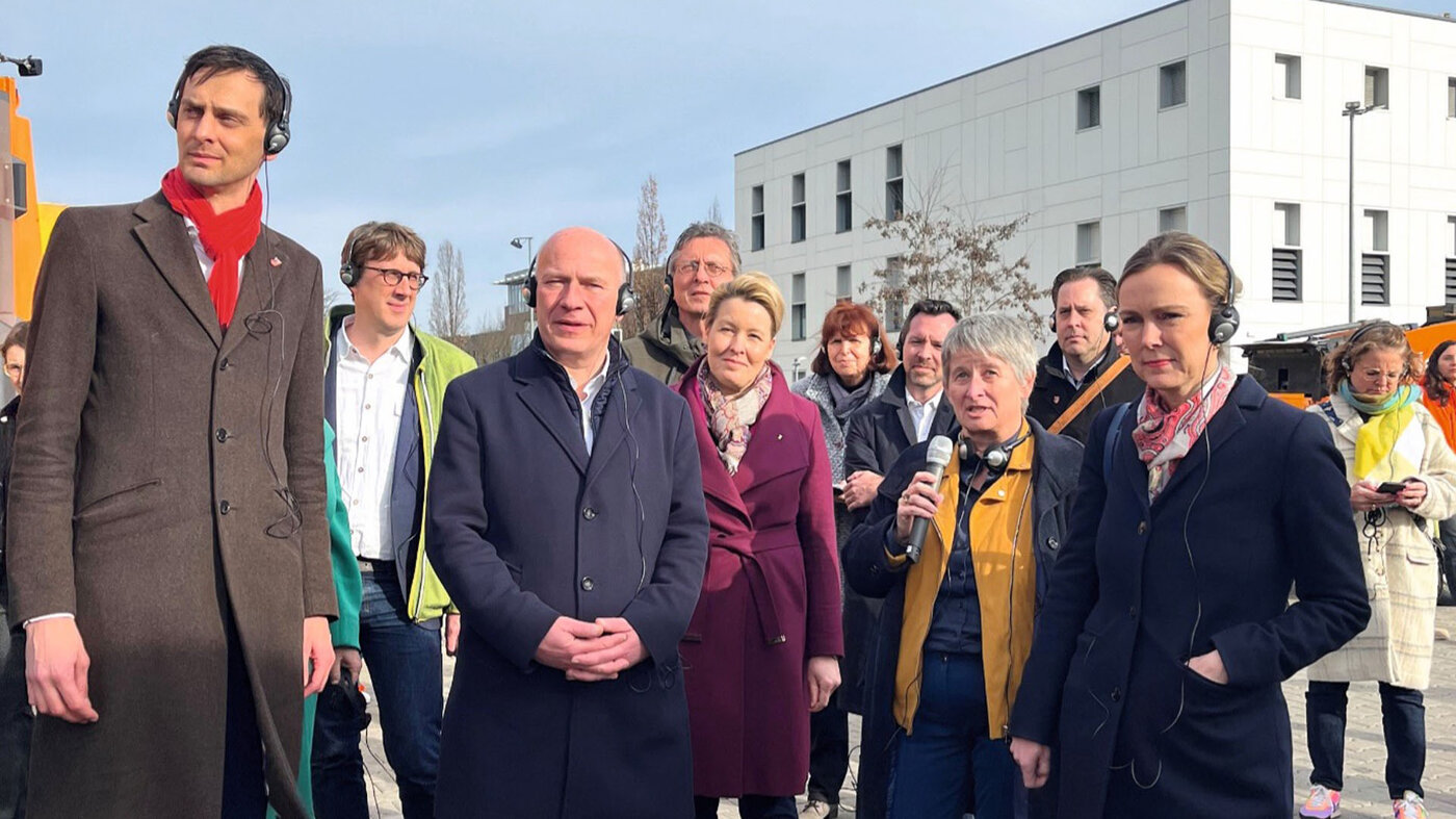 Gruppe von Politiker*innen mit Headsets steht im Freien, im Hintergrund ein modernes Gebäude bei Tageslicht.