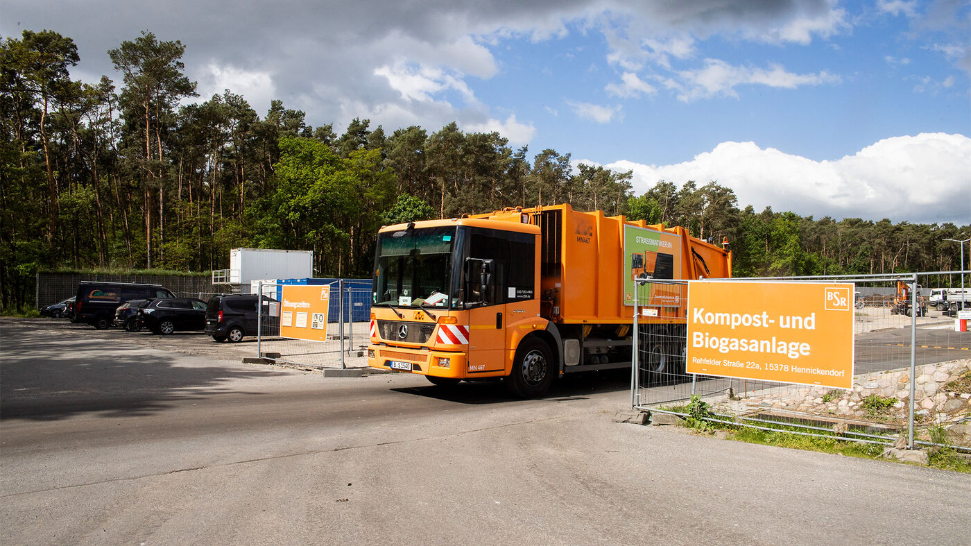 Ein orangefarbener Müllwagen fährt auf dem Gelände einer Kompost- und Biogasanlage, hinter Bäumen und einem blauen Himmel.