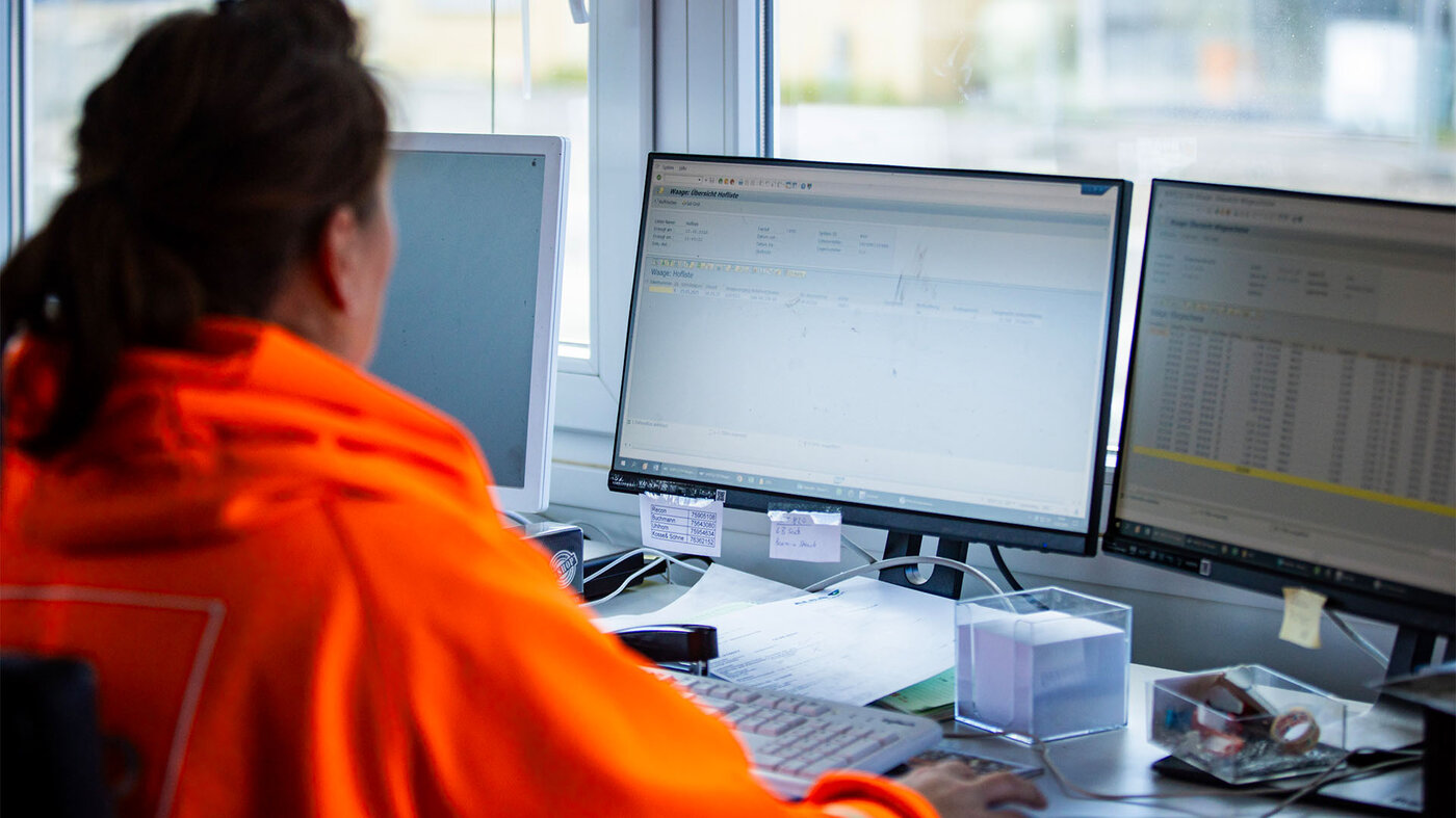 Eine BSR-Mitarbeiterin in einem leuchtend orangen Oberteil sitzt vor zwei Computermonitoren und arbeitet an einem Schreibtisch.