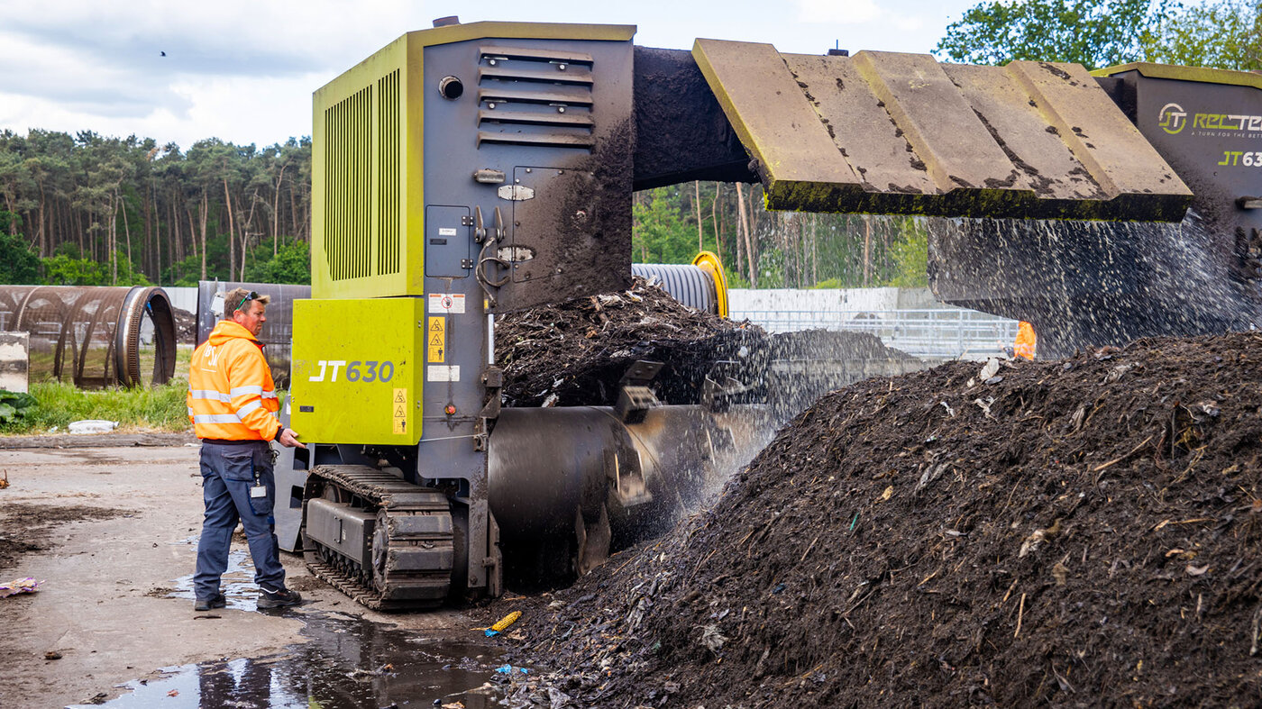 Ein Mann in Arbeitskleidung steht neben einer großen Maschine, die organisches Material zerkleinert und auf einen Haufen wirft.