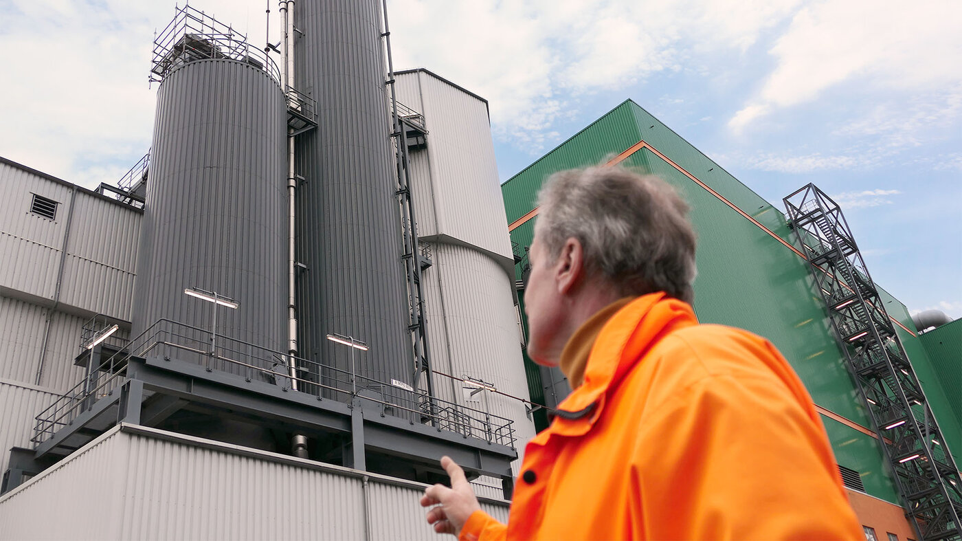 Ein Mann in orangefarbener Jacke zeigt auf industrielle Anlagen mit grauen und grünen Fassaden unter blauem Himmel.