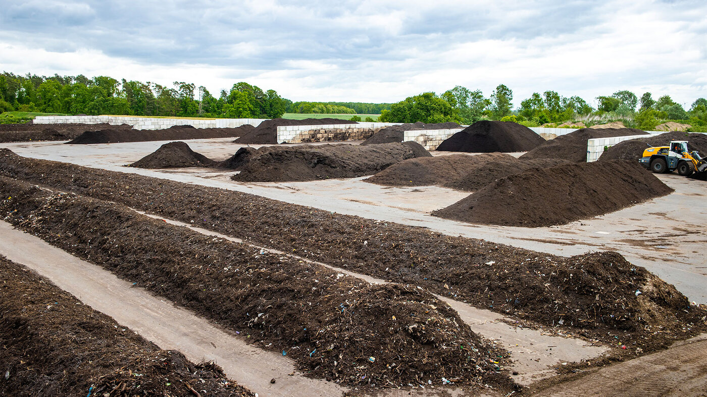 Große Kompostierungsanlage mit mehreren großen Haufen organischen Materials auf Betonflächen, umgeben von Bäumen und bewölktem Himmel.