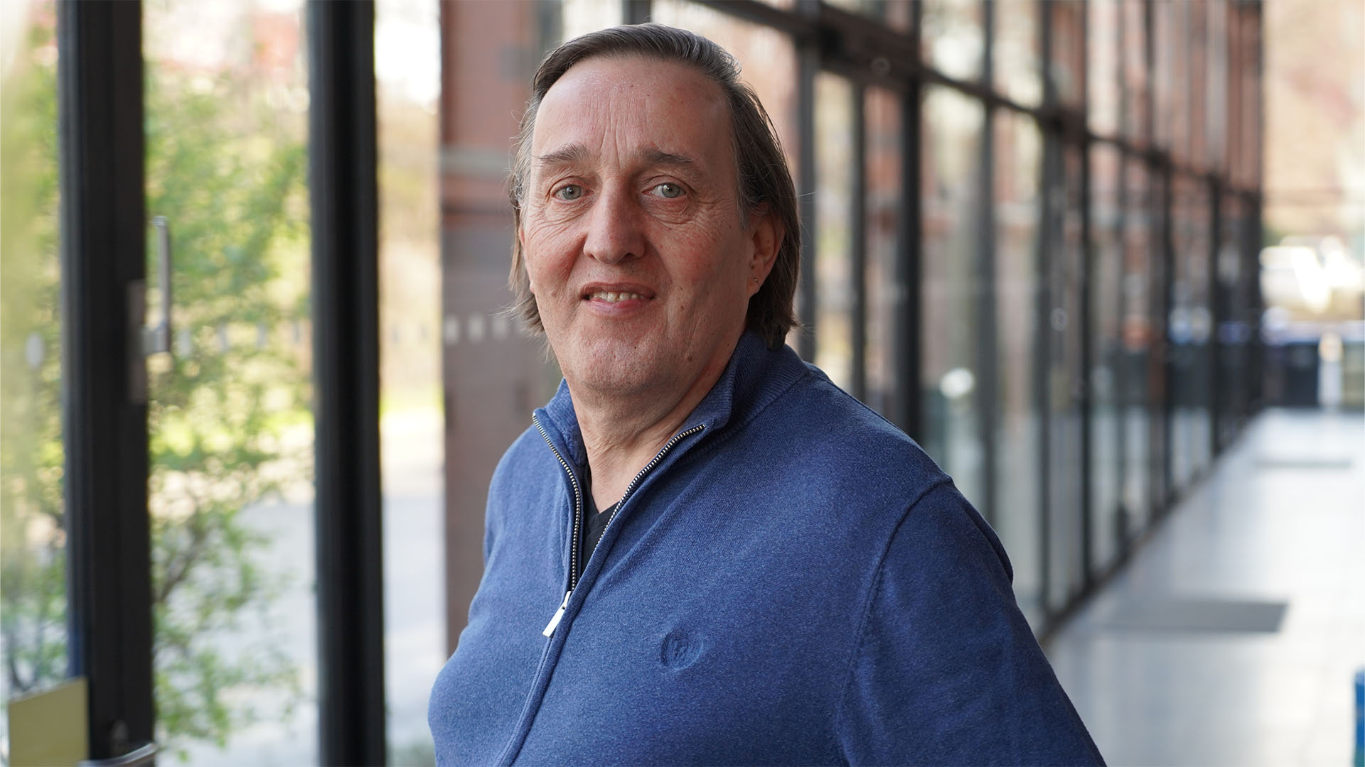 Ein Mann mit langen, dunklen Haaren und einem blauen Pullover lächelt in die Kamera. Er steht in einem modernen Gebäude mit großen Glasfenstern im Hintergrund.