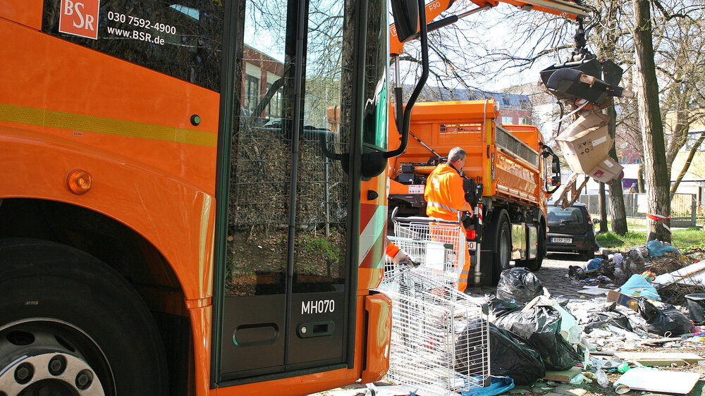 Ein orangefarbener BSR-Lastwagen mit Greifarm lädt Kartons und Müll von einem Bürgersteig in seinen Container.