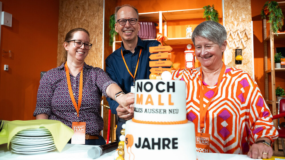Drei Personen schneiden gemeinsam eine Geburtstagstorte zum fünfjährigen Bestehen der NochMall an