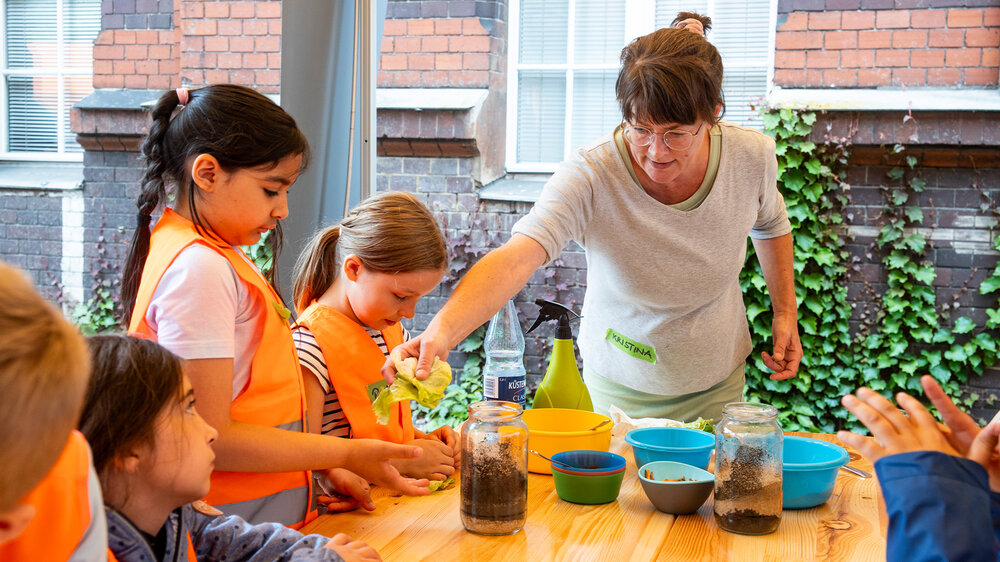 Eine Mitarbeiterin zeigt Kindern an einer Lernstation mit Erde und Blättern, wie Kompostierung funktioniert 