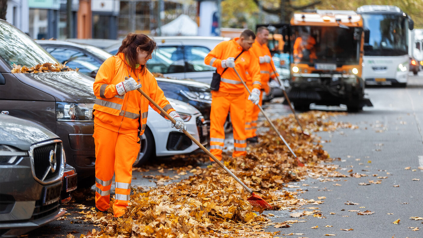 BSR-Mitarbeiter:innen kehren Laub am Straßenrand, während im Hintergrund ein Reinigungsfahrzeug im Einsatz ist