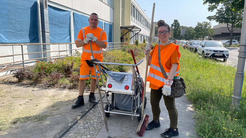 Zwei Personen in orangefarbenen Warnwesten stehen mit Reinigungsgeräten auf einem Gehweg